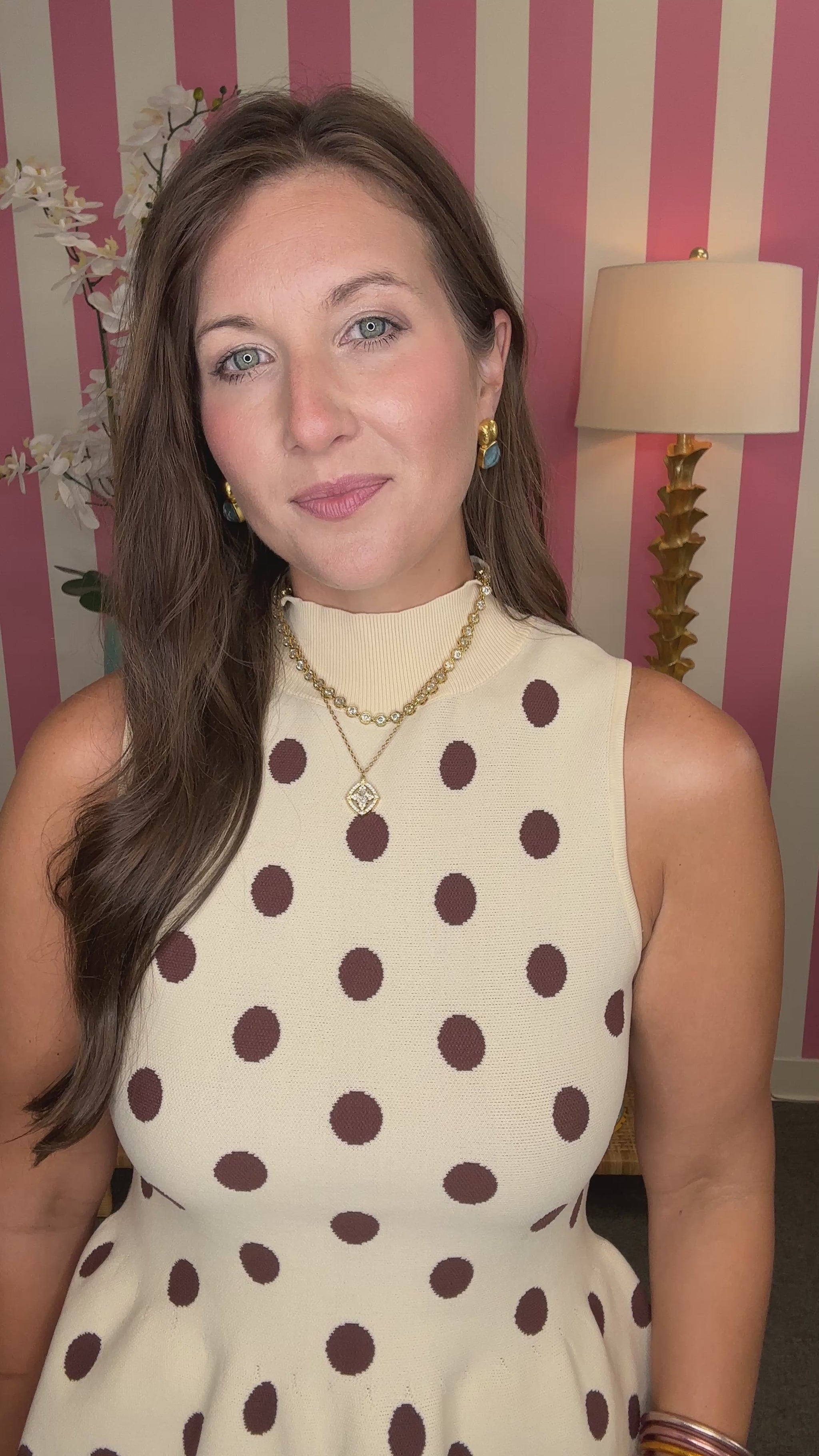A woman wearing a sleeveless cream top with brown polka dots and white pants, standing in a room with pink and white striped walls, a lamp, and a side table.

