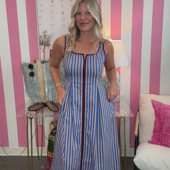 A woman modeling a sleeveless blue and white vertically striped midi dress with a front zipper, standing in front of a pink and white striped wall.