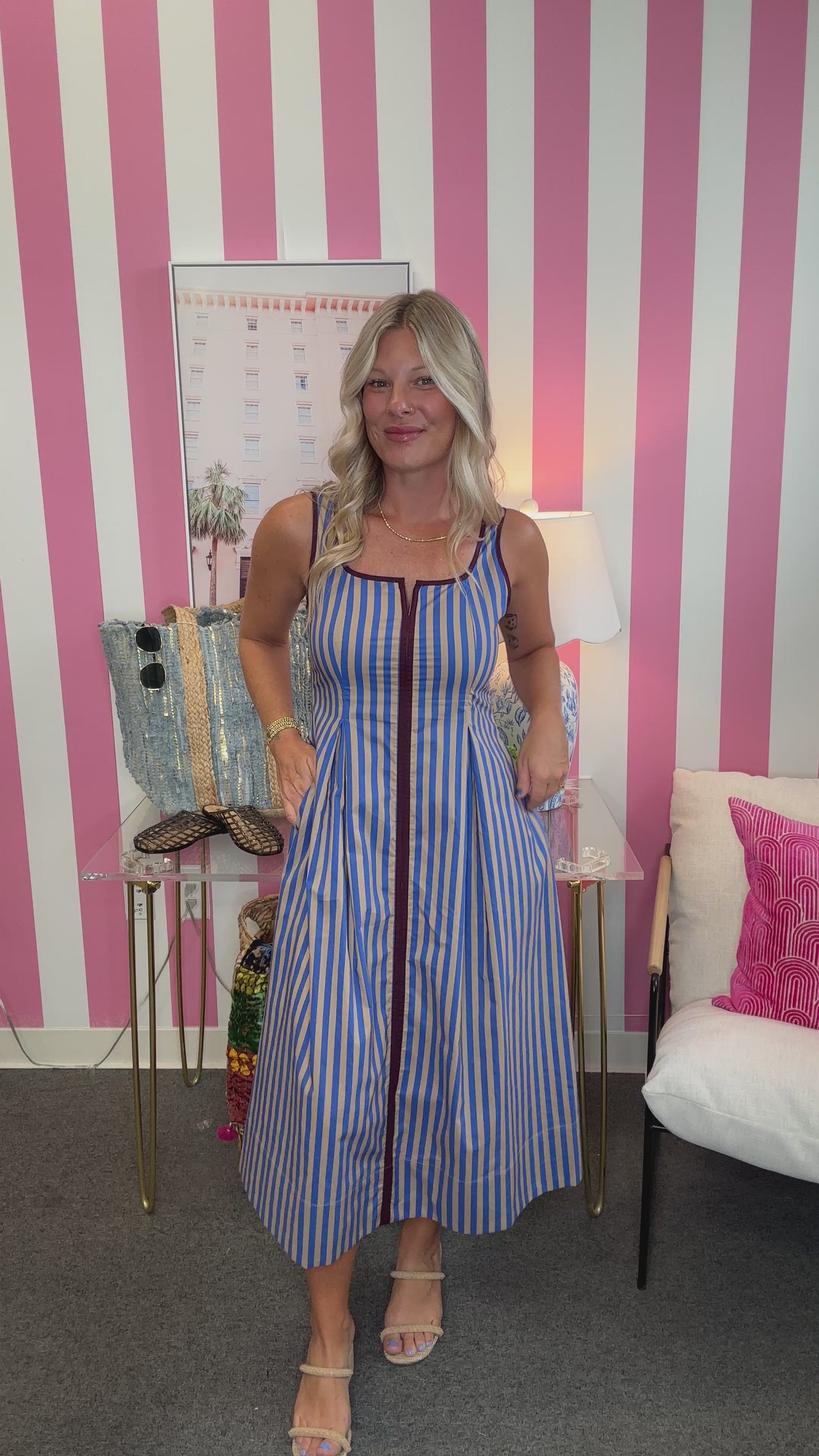 A woman modeling a sleeveless blue and white vertically striped midi dress with a front zipper, standing in front of a pink and white striped wall.
