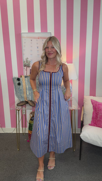 A woman modeling a sleeveless blue and white vertically striped midi dress with a front zipper, standing in front of a pink and white striped wall.