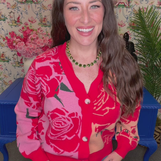 A woman wearing a red and pink cardigan with a large rose pattern, paired with blue jeans, standing in front of a floral wallpaper background.