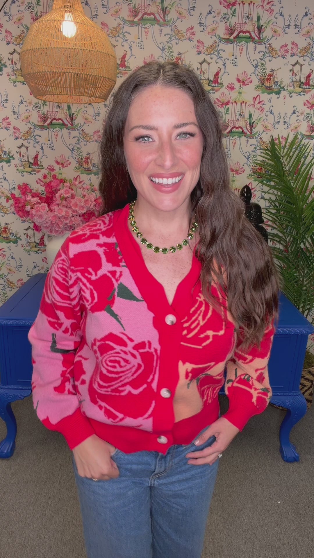 A woman wearing a red and pink cardigan with a large rose pattern, paired with blue jeans, standing in front of a floral wallpaper background.