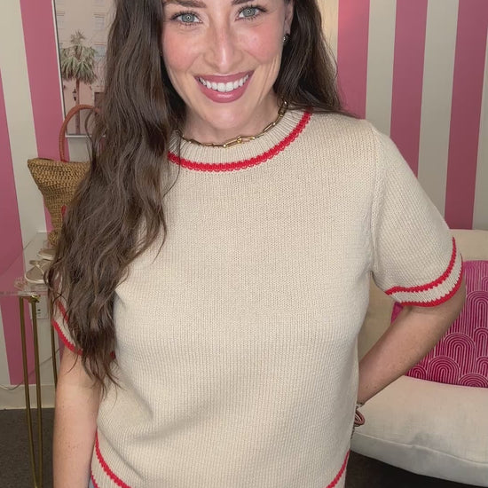 Woman wearing a beige sweater with red trim in a room with pink and white striped wallpaper.