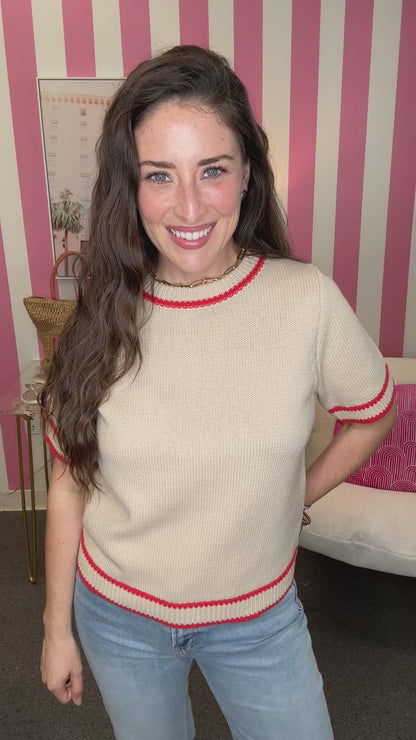 Woman wearing a beige sweater with red trim in a room with pink and white striped wallpaper.