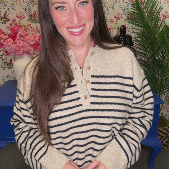 A woman with long brown hair smiling and looking over her shoulder, wearing a cream knit sweater with black wavy stripes and blue jeans, standing in a decorated room with a blue table and floral wallpaper.