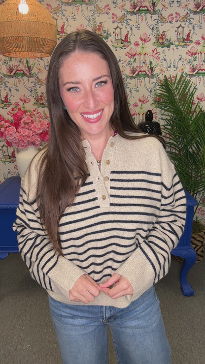 A woman with long brown hair smiling and looking over her shoulder, wearing a cream knit sweater with black wavy stripes and blue jeans, standing in a decorated room with a blue table and floral wallpaper.