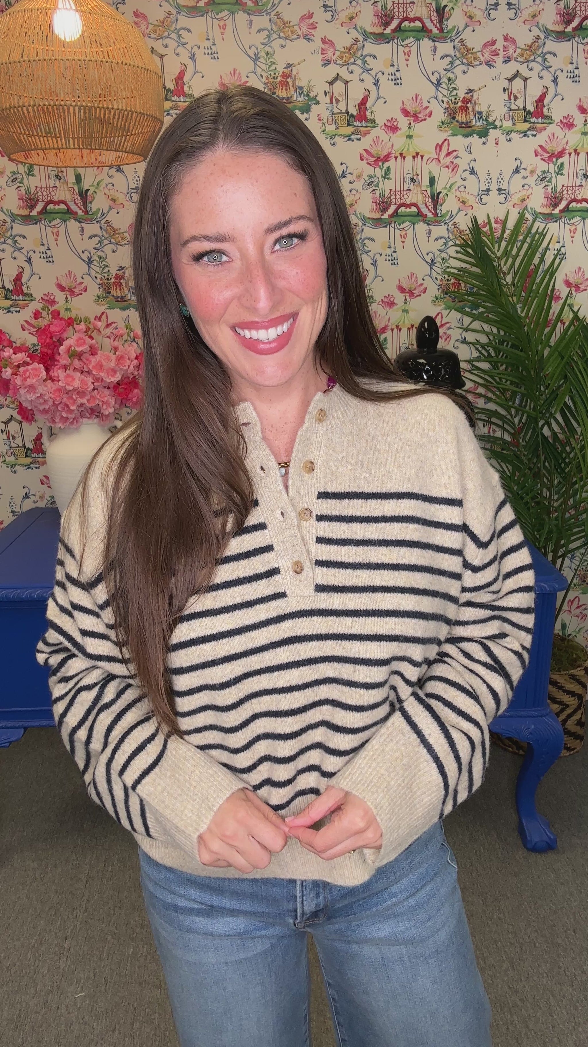 A woman with long brown hair smiling and looking over her shoulder, wearing a cream knit sweater with black wavy stripes and blue jeans, standing in a decorated room with a blue table and floral wallpaper.