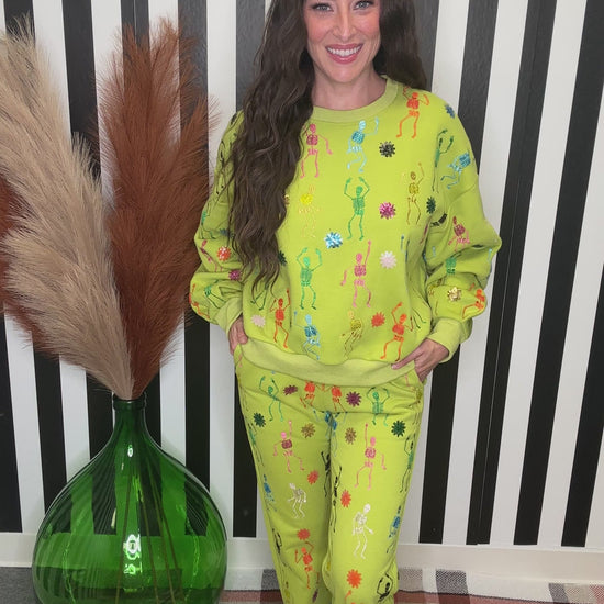A woman modeling a bright yellow-green matching lounge set with colorful floral prints, standing in front of a black and white striped wall next to a green glass vase with pampas grass.