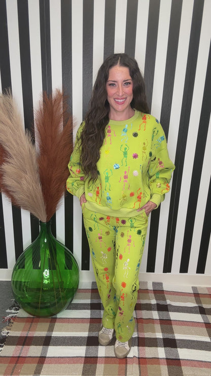 A woman modeling a bright yellow-green matching lounge set with colorful floral prints, standing in front of a black and white striped wall next to a green glass vase with pampas grass.