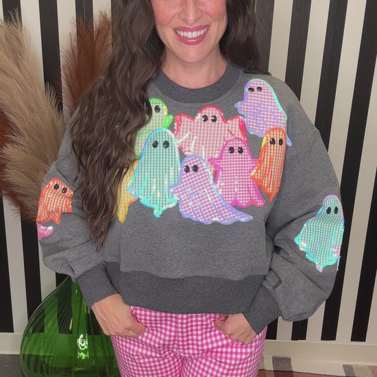 A woman wearing a gray crewneck sweatshirt with colorful gingham ghosts on the front and pink gingham pants, standing in front of a black and white striped background.