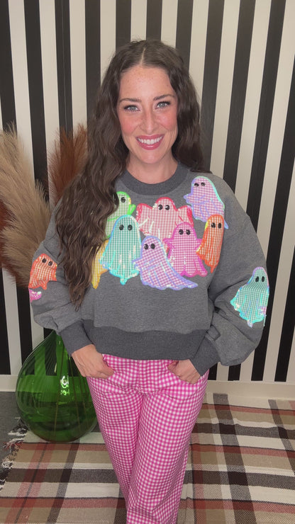 A woman wearing a gray crewneck sweatshirt with colorful gingham ghosts on the front and pink gingham pants, standing in front of a black and white striped background.
