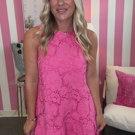 A woman wearing a bright pink lace dress with a large bow on the back, shown from behind in a room with pink and white striped walls.