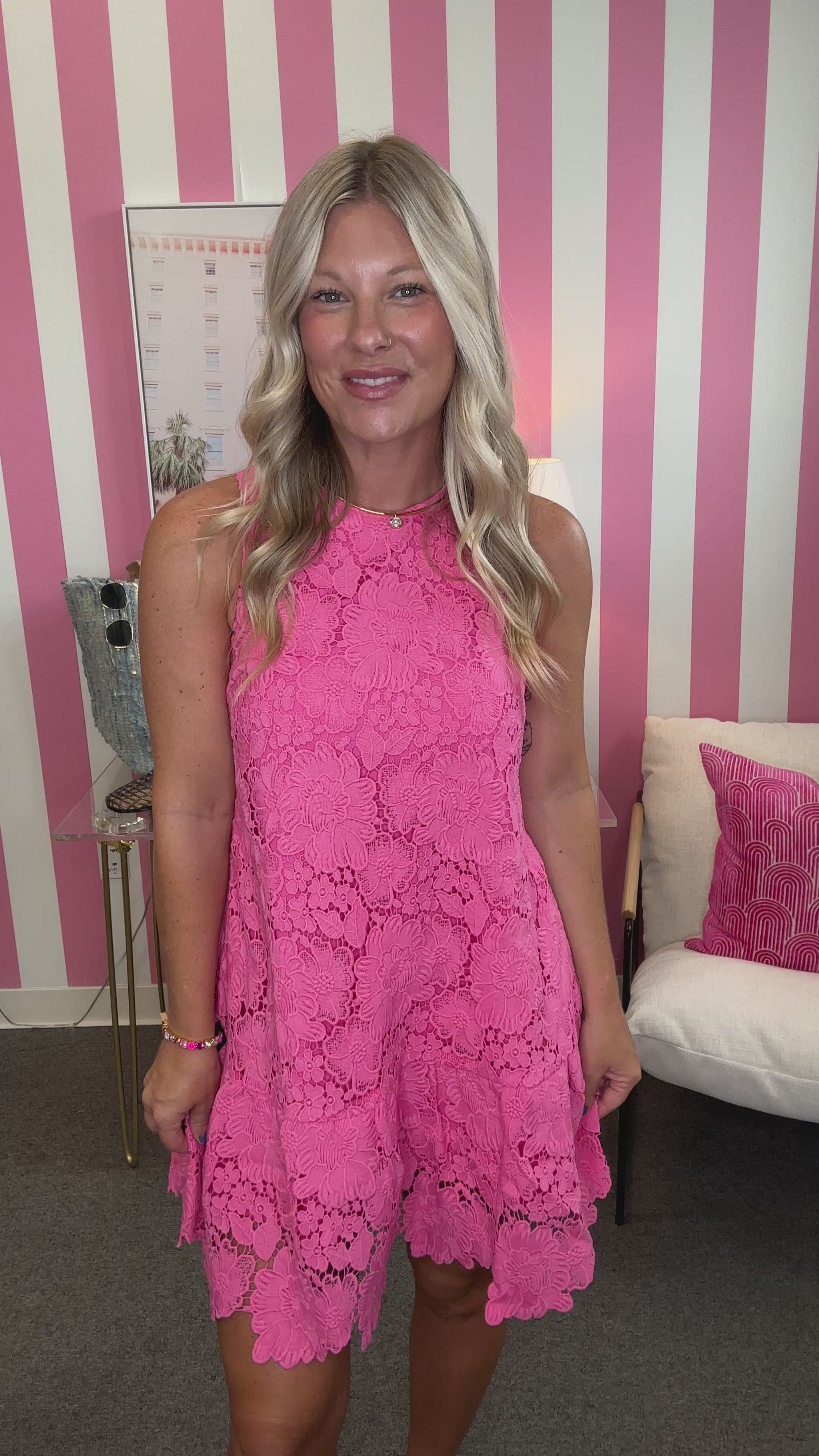 A woman wearing a bright pink lace dress with a large bow on the back, shown from behind in a room with pink and white striped walls.