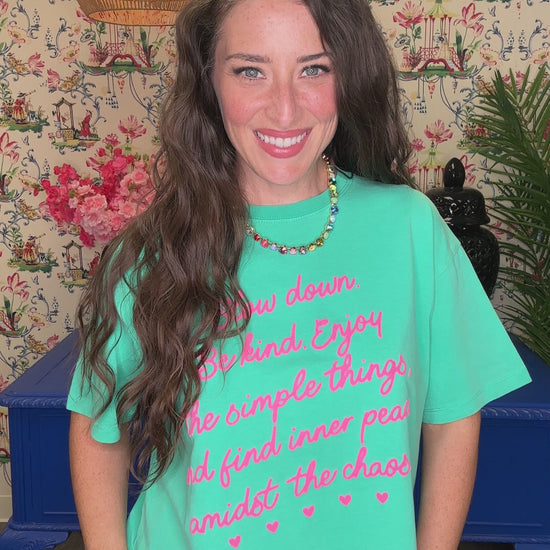 A woman smiling and wearing a turquoise graphic t-shirt with pink text that reads 'Slow down. Be kind.