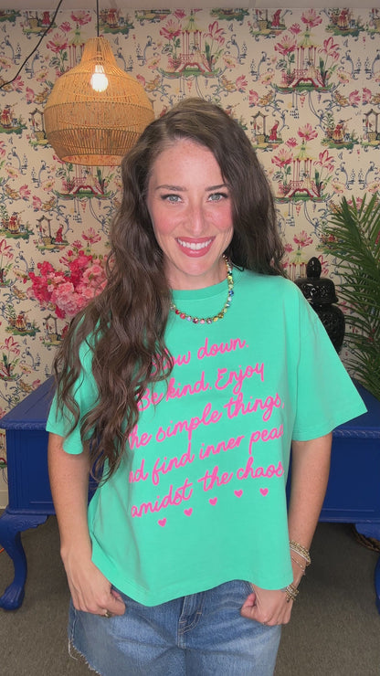 A woman smiling and wearing a turquoise graphic t-shirt with pink text that reads 'Slow down. Be kind.