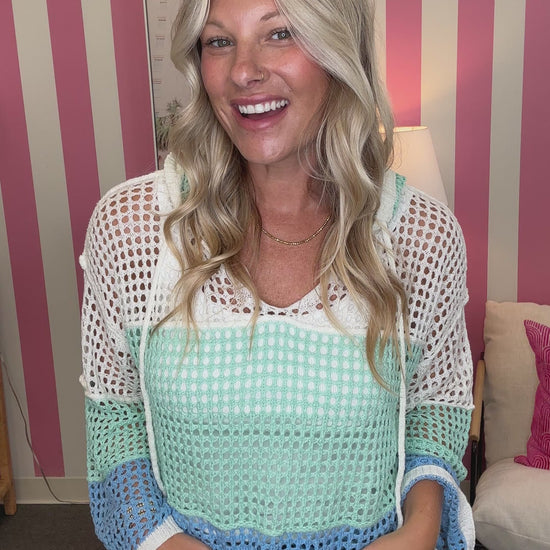 A smiling blonde woman wearing a colorful crochet knit top with white, mint green, and light blue sections, paired with blue jeans, standing in front of a pink and white striped wall.