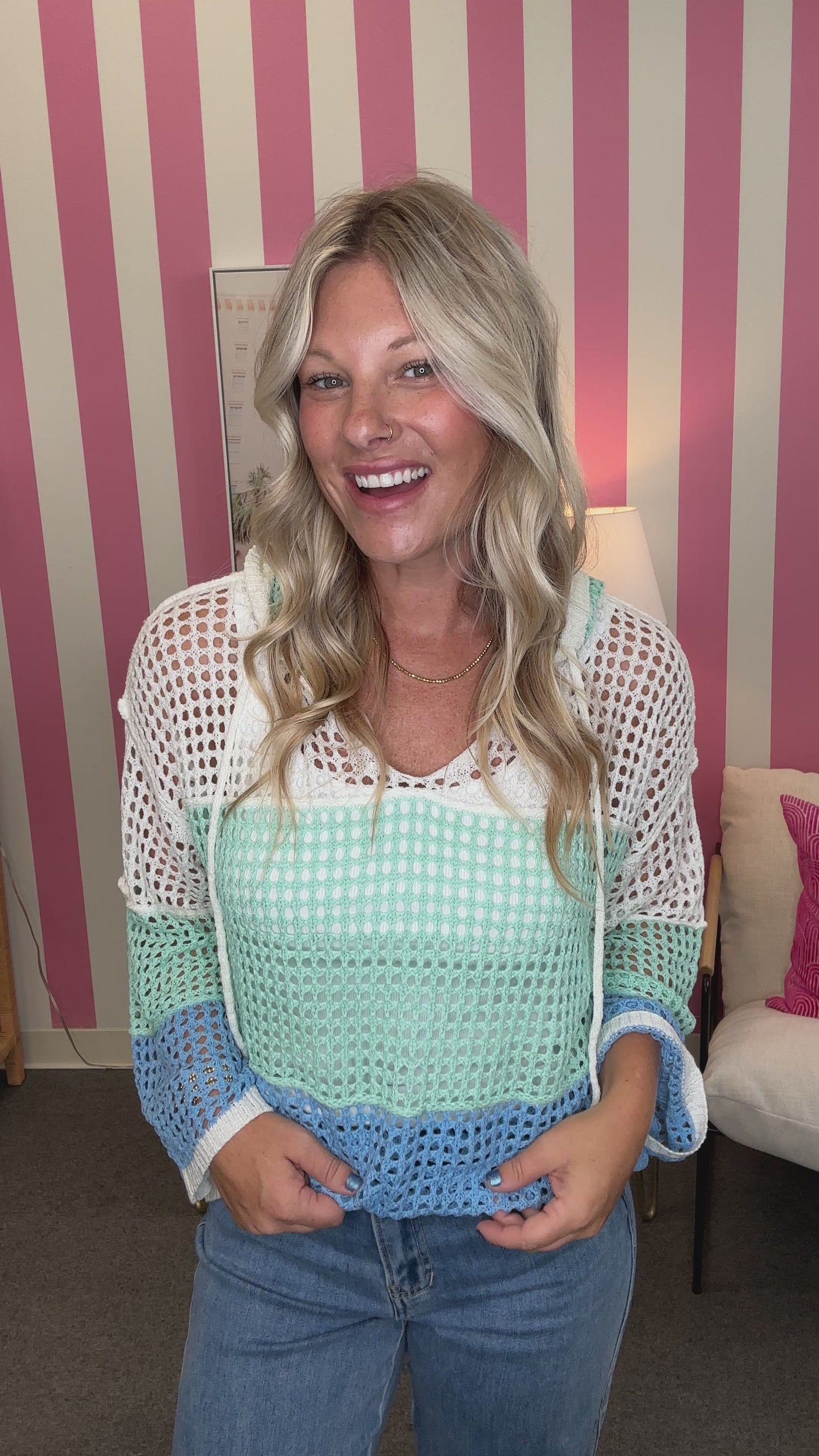 A smiling blonde woman wearing a colorful crochet knit top with white, mint green, and light blue sections, paired with blue jeans, standing in front of a pink and white striped wall.
