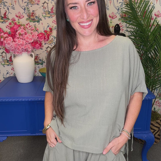 A woman modeling a sage green short-sleeve romper with a relaxed fit, standing in front of a floral wallpaper background with a blue bench.