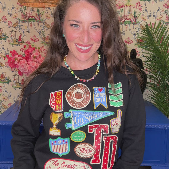 A smiling woman with long brown hair wears a black long-sleeve sweatshirt covered in colorful football-themed patches and graphics, including 'TD', 'Peanut Butter', 'Cheddar', and 'The Great American Football Crawl'. She is standing indoors against a floral wallpaper background with a blue table visible behind her.