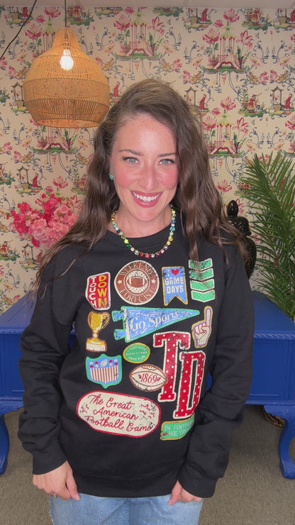 A smiling woman with long brown hair wears a black long-sleeve sweatshirt covered in colorful football-themed patches and graphics, including 'TD', 'Peanut Butter', 'Cheddar', and 'The Great American Football Crawl'. She is standing indoors against a floral wallpaper background with a blue table visible behind her.