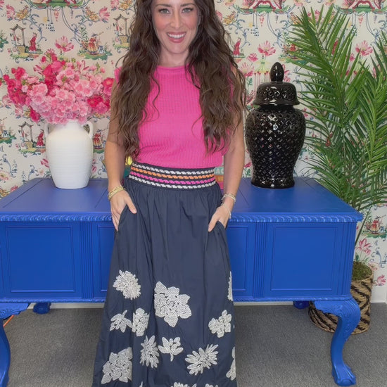 A woman modeling a sleeveless pink ribbed knit top paired with a navy blue maxi skirt featuring white floral embroidery and a striped waistband, posed in front of a blue cabinet and floral wallpaper.