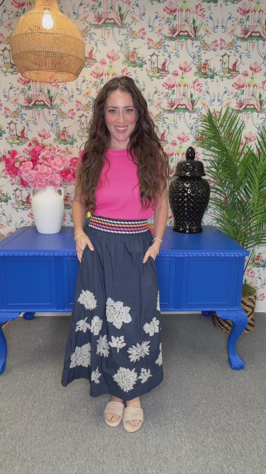 A woman modeling a sleeveless pink ribbed knit top paired with a navy blue maxi skirt featuring white floral embroidery and a striped waistband, posed in front of a blue cabinet and floral wallpaper.