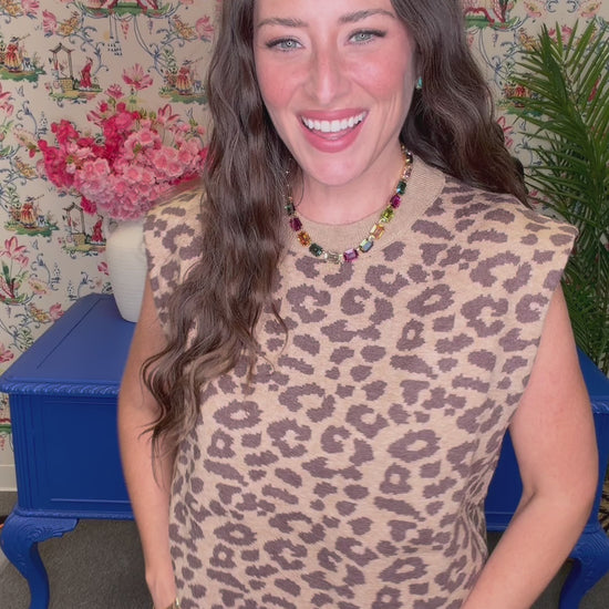 A woman with long wavy brown hair smiling, wearing a sleeveless beige knit top with a brown leopard print pattern and a colorful beaded necklace. She is standing in front of a floral wallpaper background with blue furniture visible.