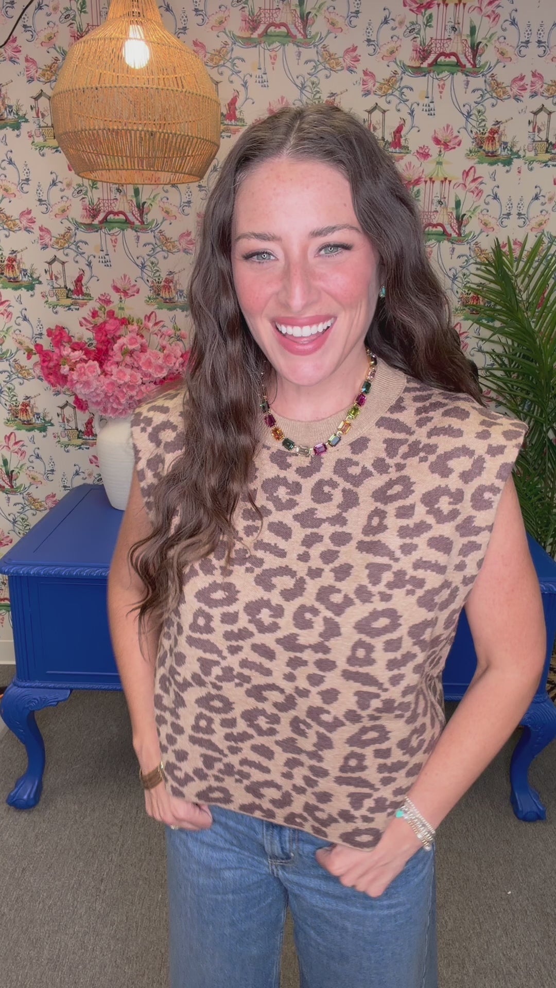 A woman with long wavy brown hair smiling, wearing a sleeveless beige knit top with a brown leopard print pattern and a colorful beaded necklace. She is standing in front of a floral wallpaper background with blue furniture visible.