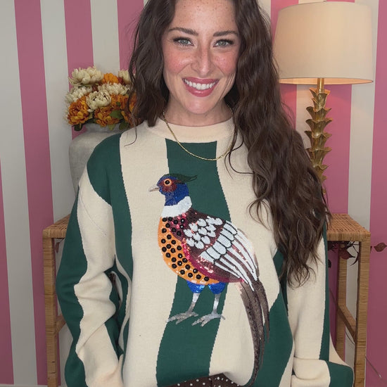 A woman wearing a green and cream striped sweater with a colorful pheasant bird graphic on the front, standing in front of a pink and white striped wall.
