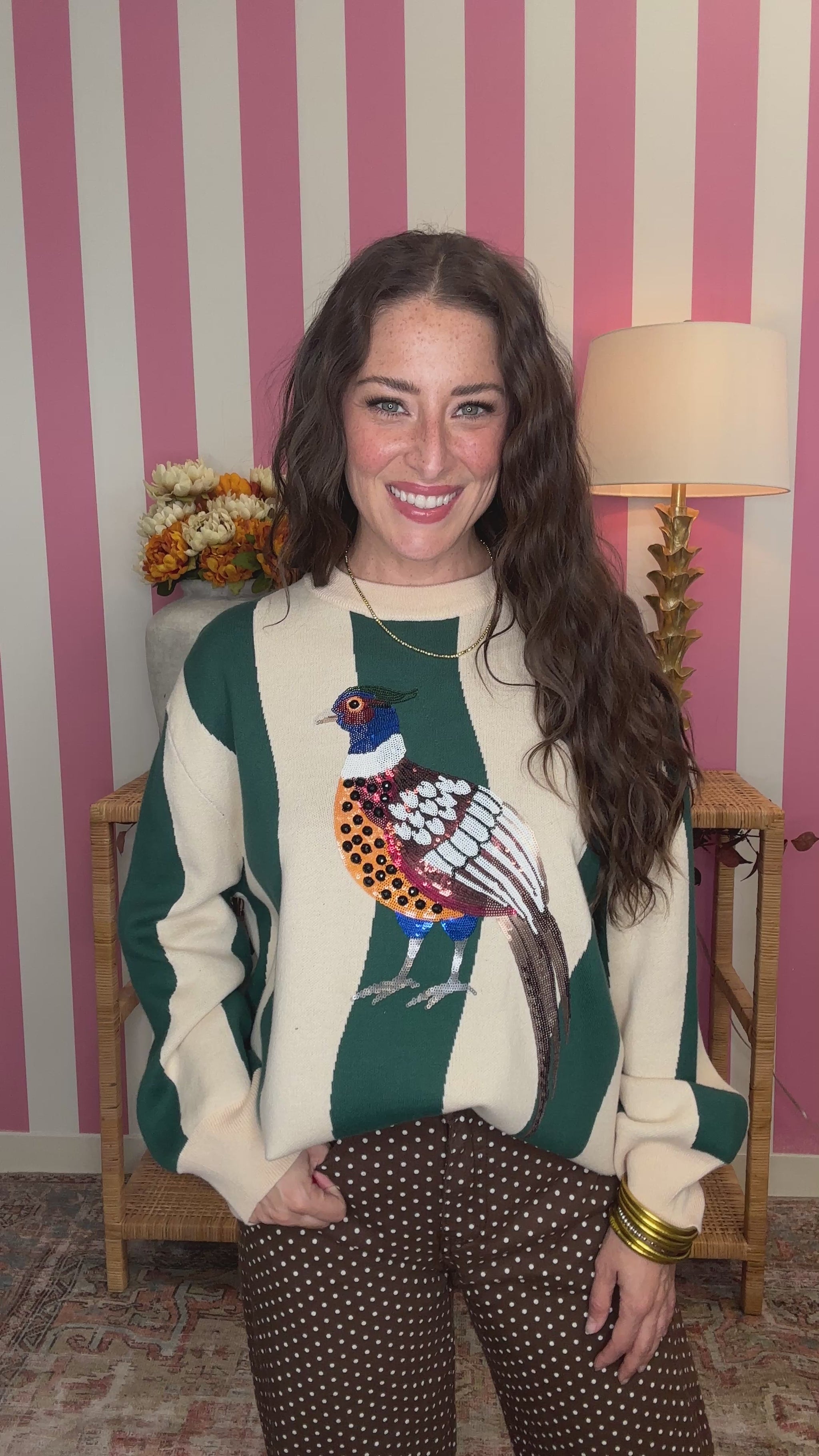 A woman wearing a green and cream striped sweater with a colorful pheasant bird graphic on the front, standing in front of a pink and white striped wall.