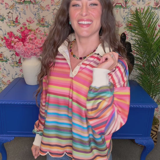 A woman with long wavy brown hair smiles while wearing a colorful striped long-sleeve blouse with a scalloped hem, standing in a decorated room with floral wallpaper, a blue side table, and plants.