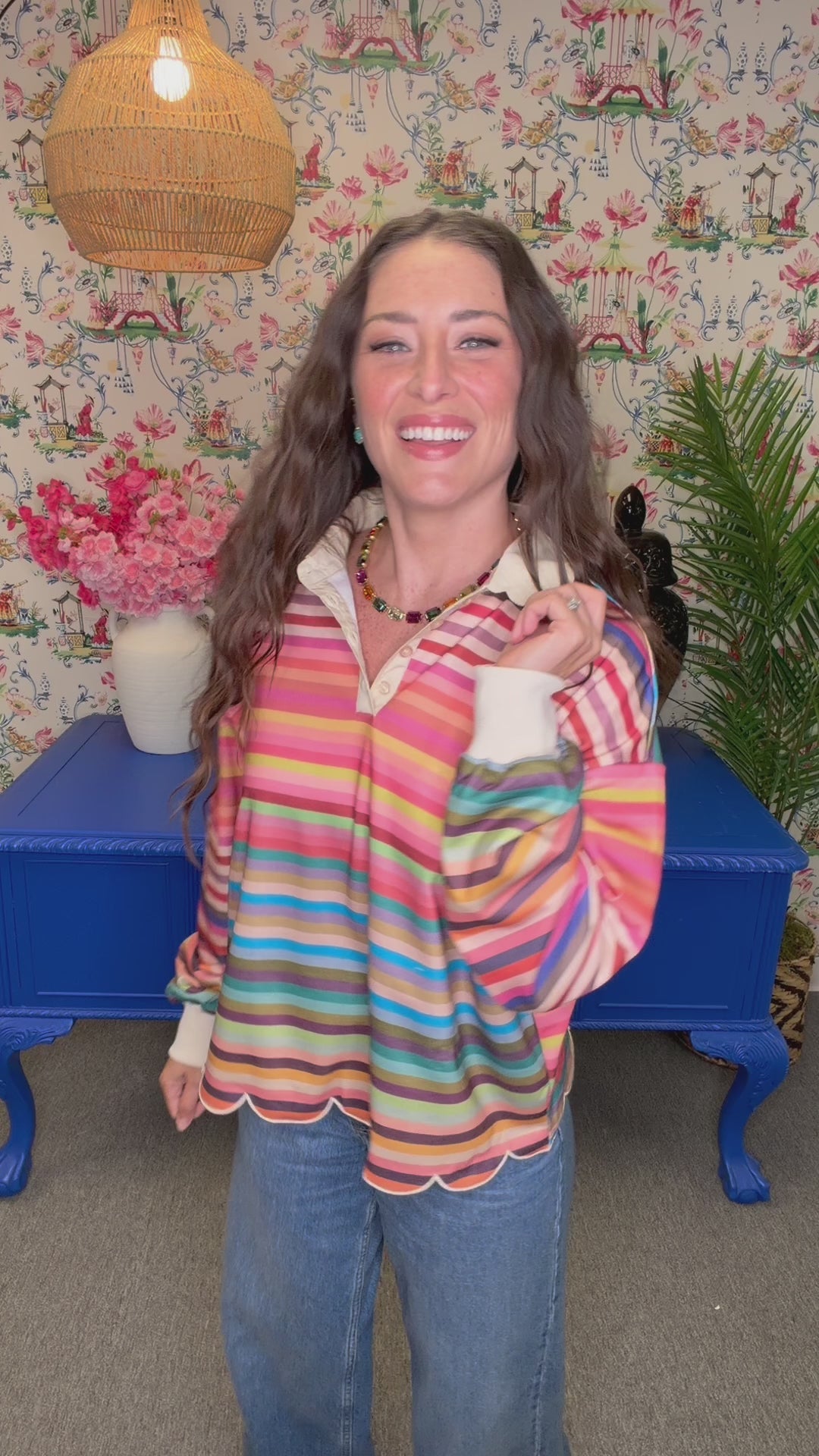 A woman with long wavy brown hair smiles while wearing a colorful striped long-sleeve blouse with a scalloped hem, standing in a decorated room with floral wallpaper, a blue side table, and plants.