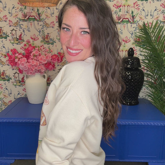 A woman smiling while wearing a cream-colored crewneck sweatshirt with a colorful graphic print that reads 'It's a Good Day For Bape' surrounded by sports and coffee-themed illustrations. The background features a floral wallpaper, a hanging lamp, and blue furniture.