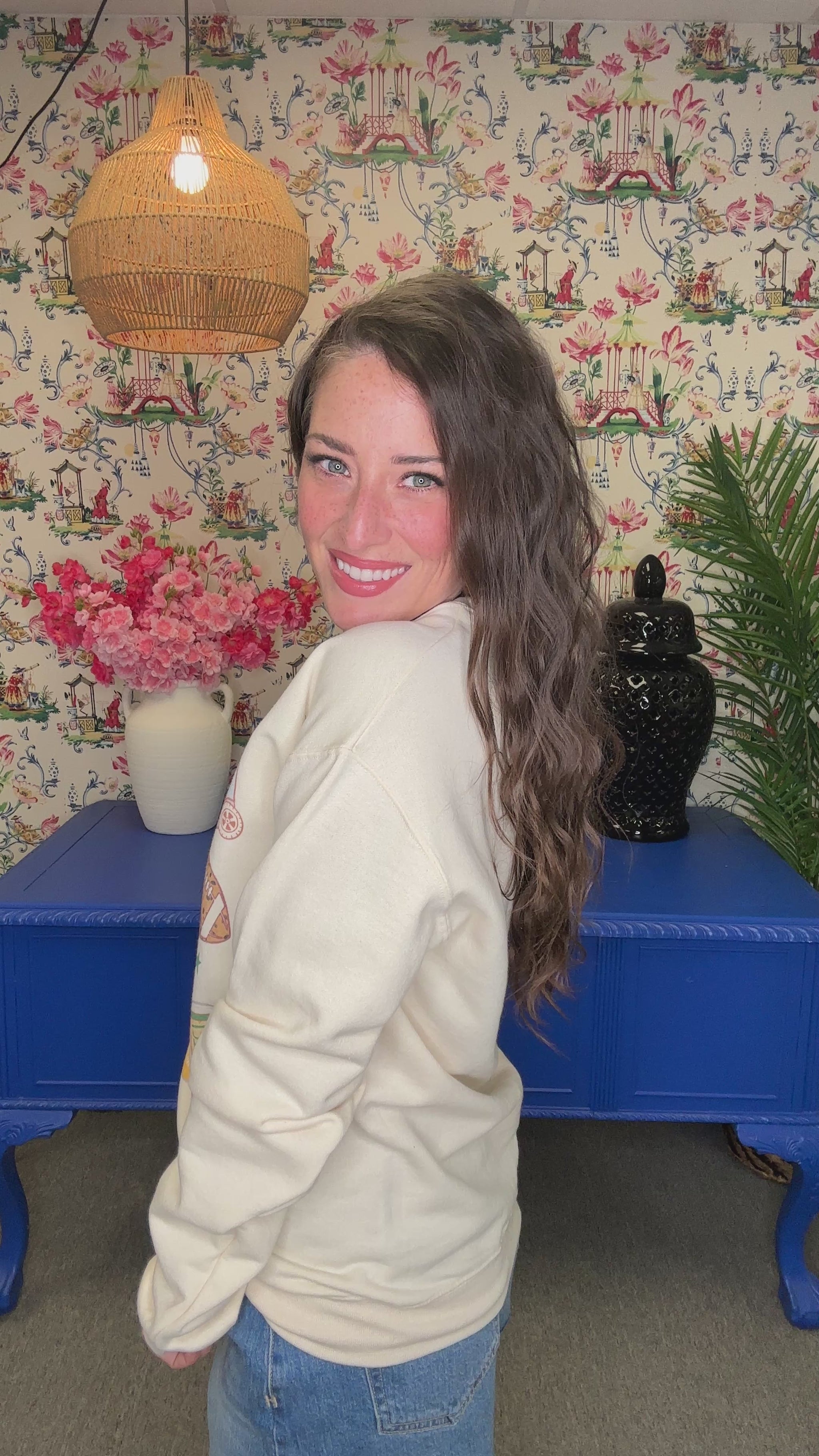 A woman smiling while wearing a cream-colored crewneck sweatshirt with a colorful graphic print that reads 'It's a Good Day For Bape' surrounded by sports and coffee-themed illustrations. The background features a floral wallpaper, a hanging lamp, and blue furniture.