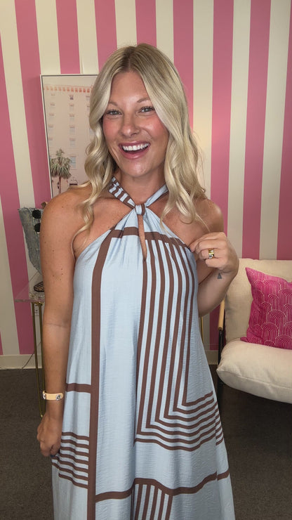 A woman modeling a light blue sleeveless maxi dress with brown geometric stripe patterns, standing in a room with pink and white striped walls.