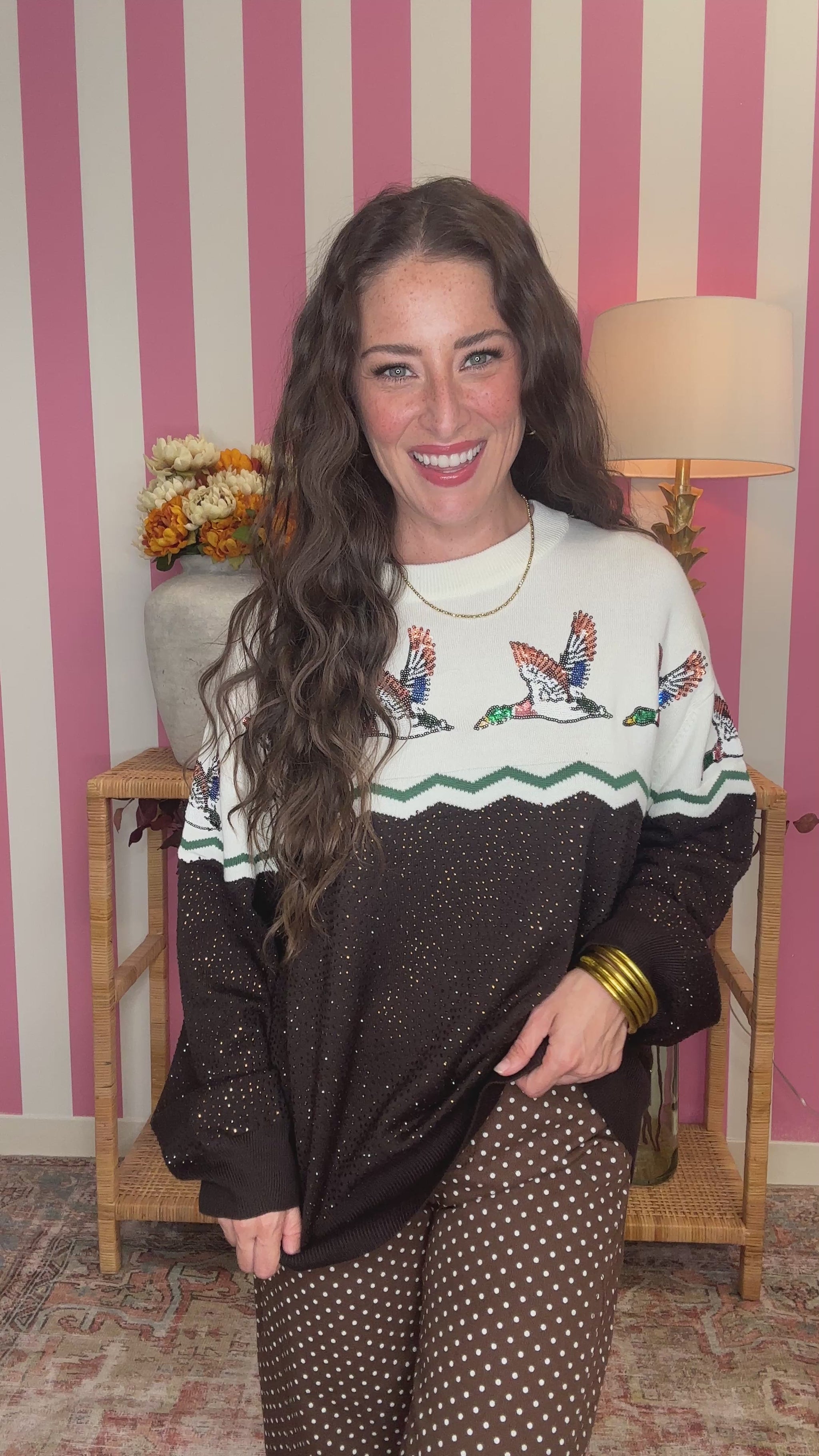 A woman wearing a long-sleeve knit sweater with a white upper section featuring embroidered ducks, a green zigzag stripe, and a black lower section with small gold-tone dot details. She is posing in front of a pink and white striped wall.