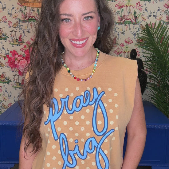 A smiling woman wearing a tan sleeveless crop top with blue script reading 'pray big' and white polka dots, paired with blue jeans, in a decorated indoor setting.