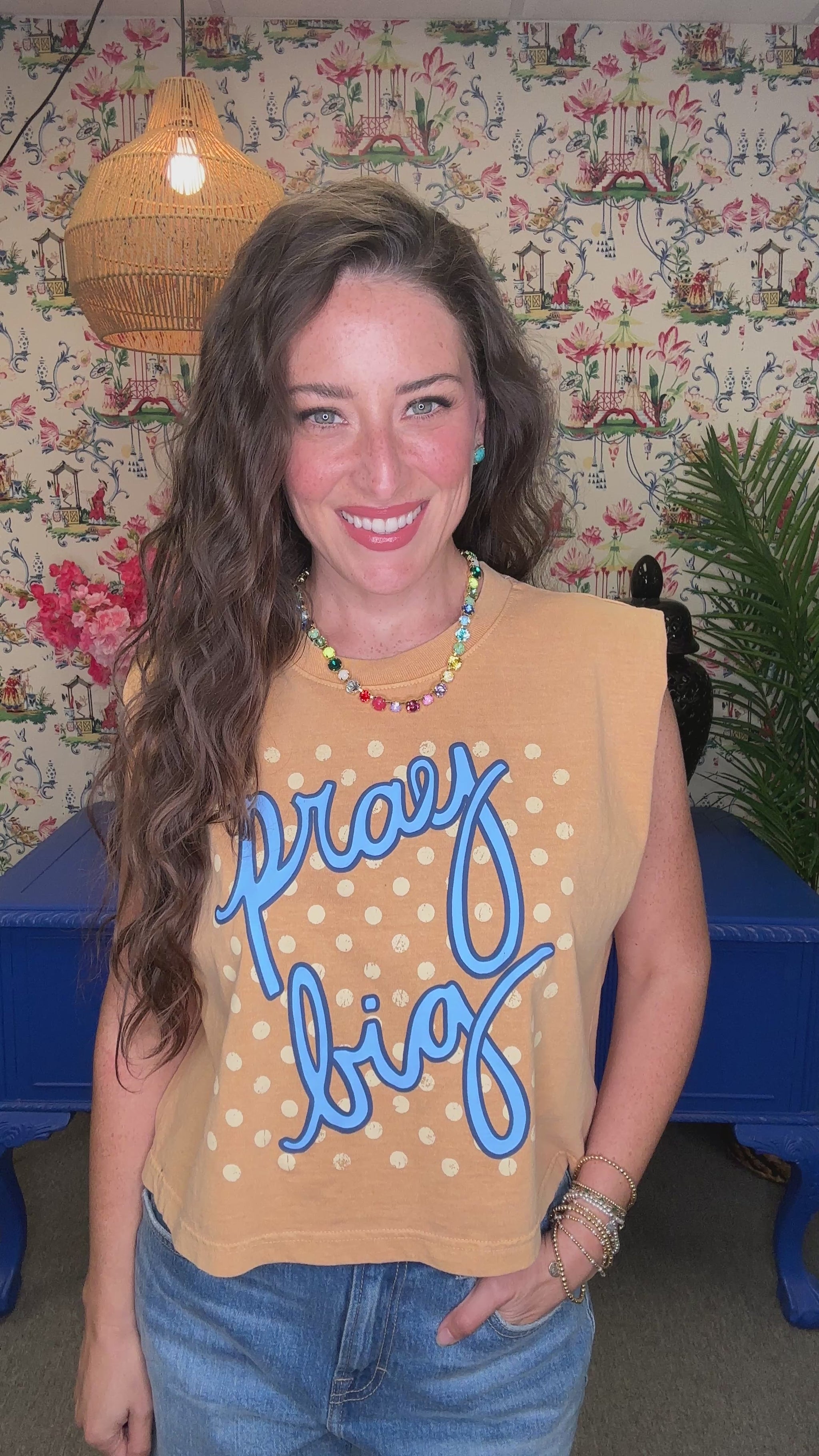 A smiling woman wearing a tan sleeveless crop top with blue script reading 'pray big' and white polka dots, paired with blue jeans, in a decorated indoor setting.
