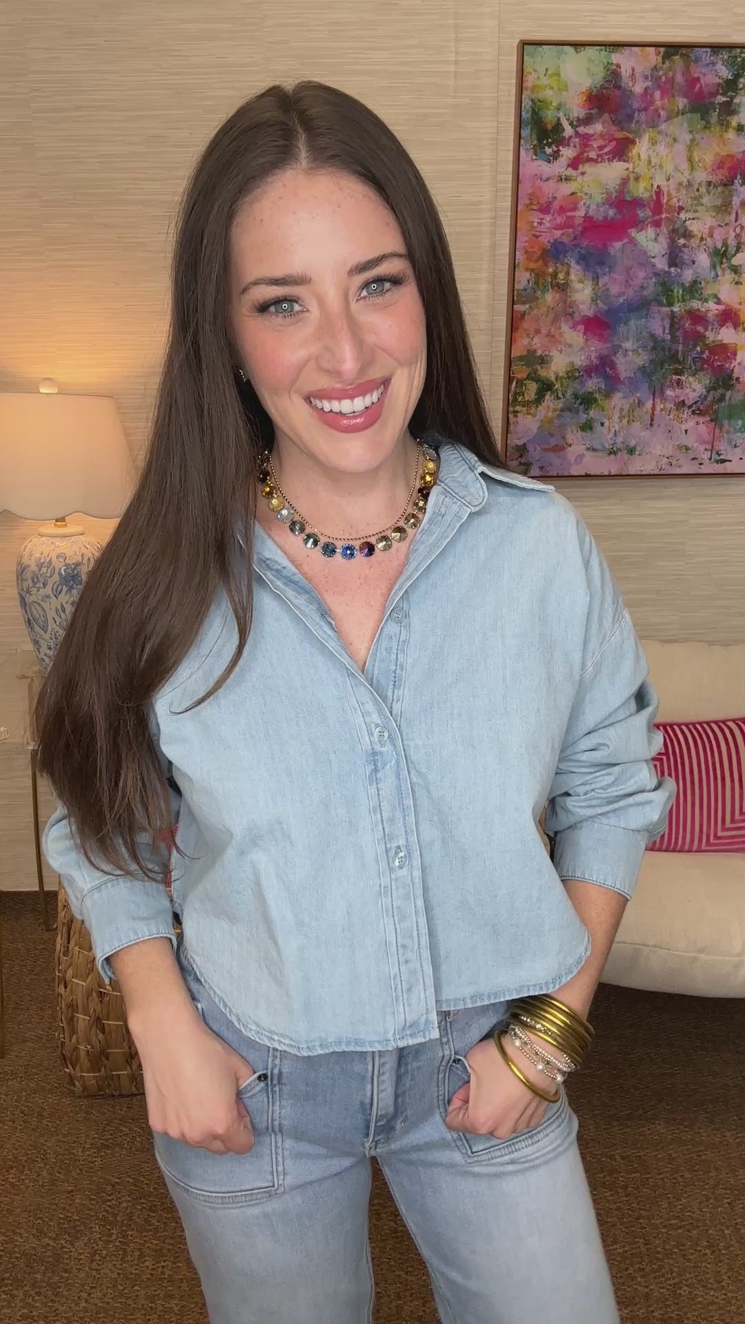Woman wearing a light blue denim shirt and jeans in a room with a colorful painting on the wall.