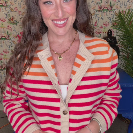 A woman smiling and wearing a cream cardigan with bold pink and orange horizontal stripes, paired with blue jeans, in a retro-themed room with floral wallpaper and a blue bench.