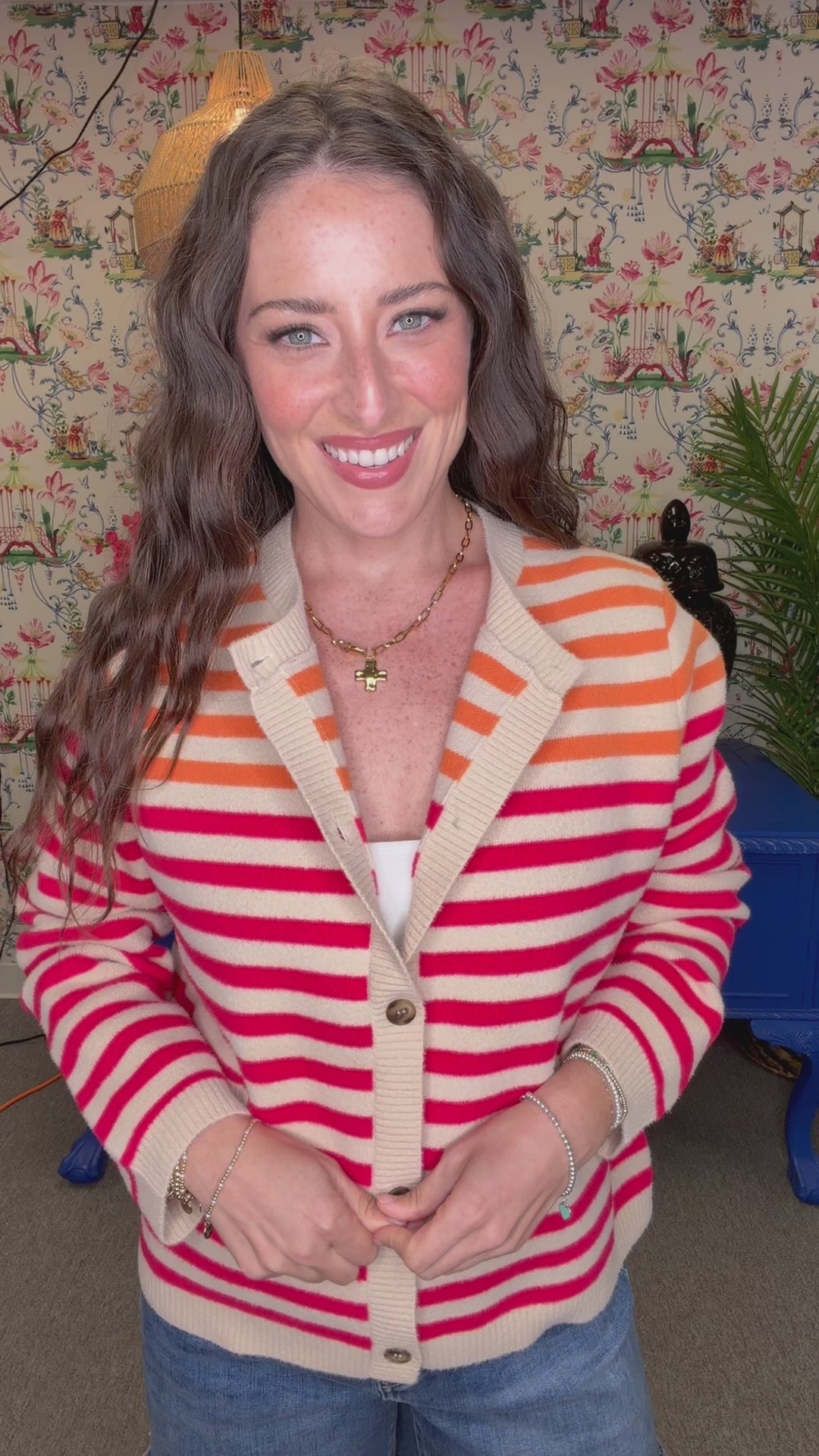 A woman smiling and wearing a cream cardigan with bold pink and orange horizontal stripes, paired with blue jeans, in a retro-themed room with floral wallpaper and a blue bench.