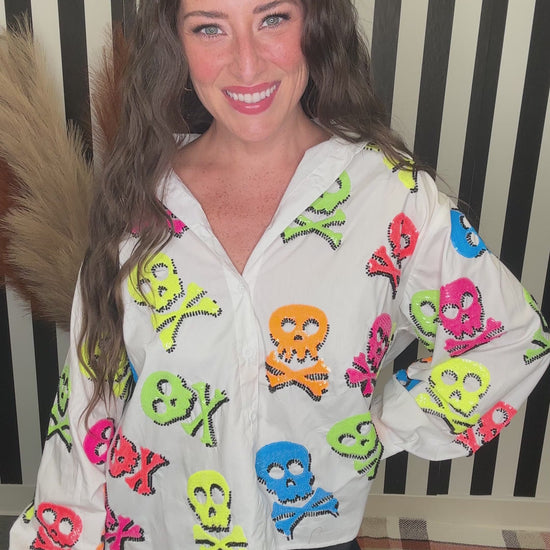 A woman smiling while wearing a white button-up shirt covered in colorful skull and crossbones motifs, paired with black pants, standing in front of a black and white striped background.