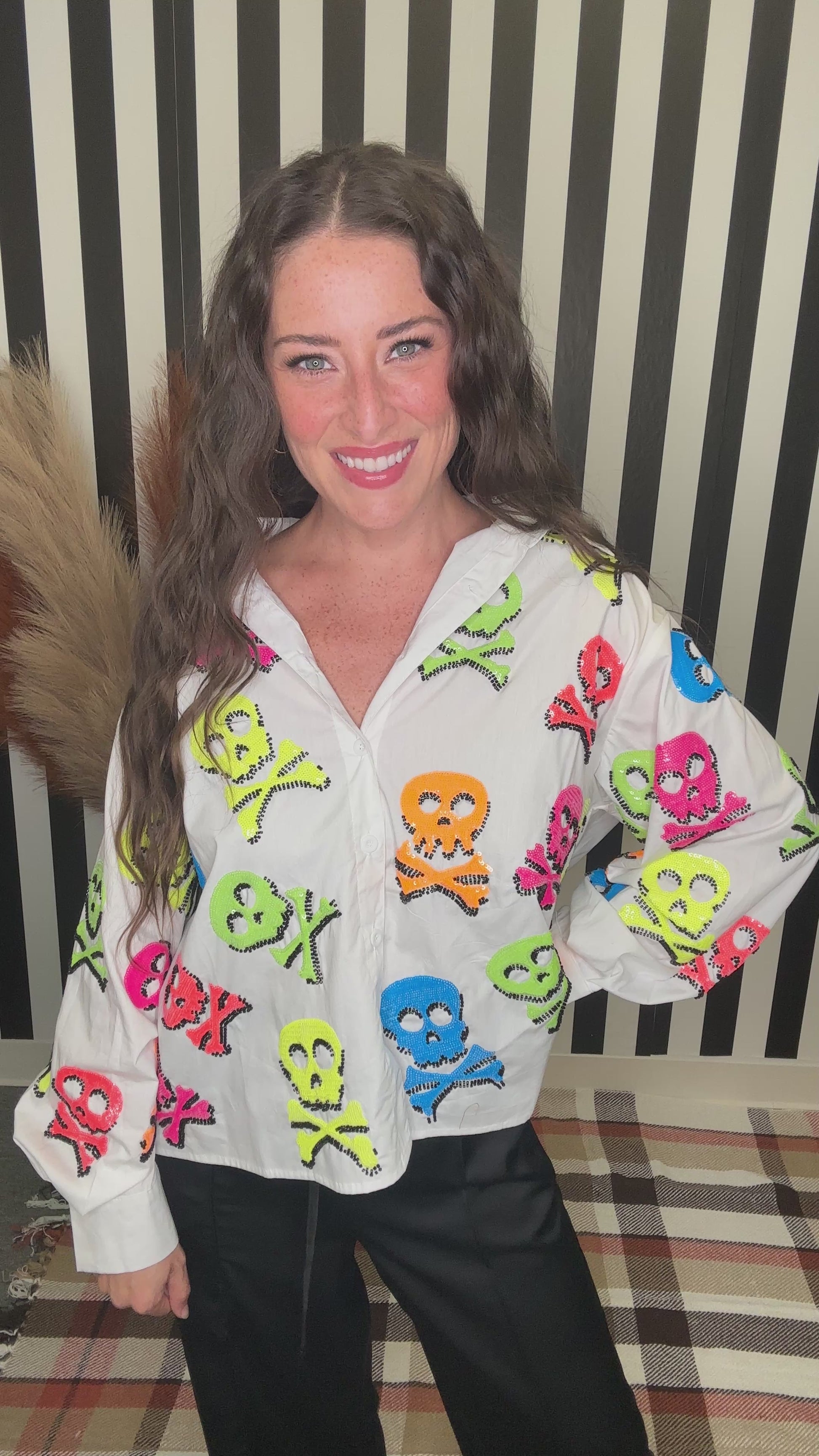 A woman smiling while wearing a white button-up shirt covered in colorful skull and crossbones motifs, paired with black pants, standing in front of a black and white striped background.