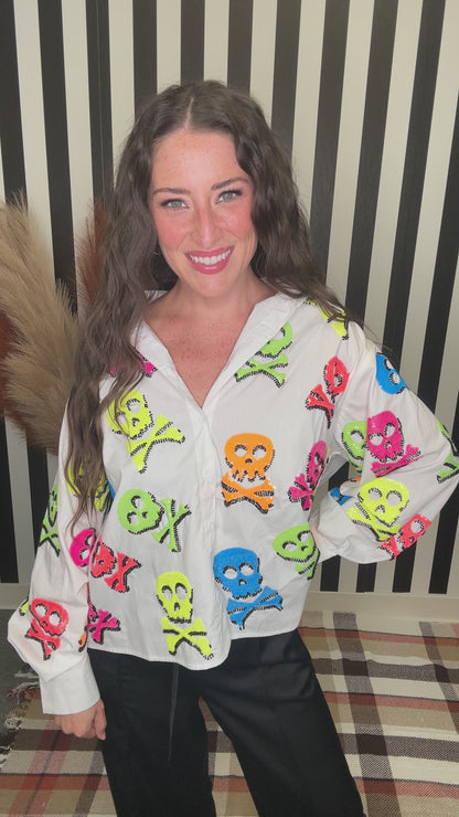 A woman smiling while wearing a white button-up shirt covered in colorful skull and crossbones motifs, paired with black pants, standing in front of a black and white striped background.