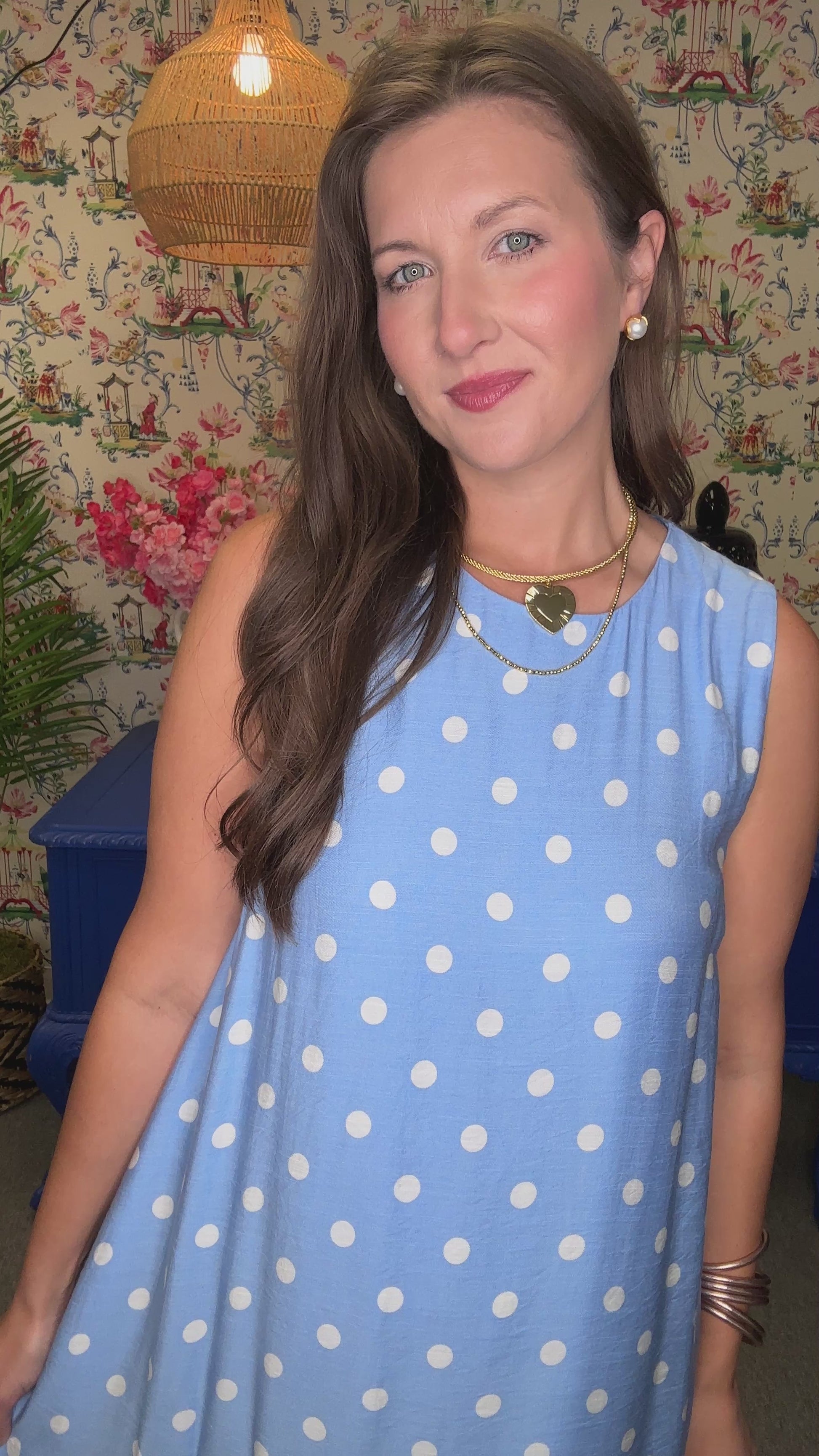 Woman in a blue polka dot dress standing in a room with floral wallpaper and a blue bench.