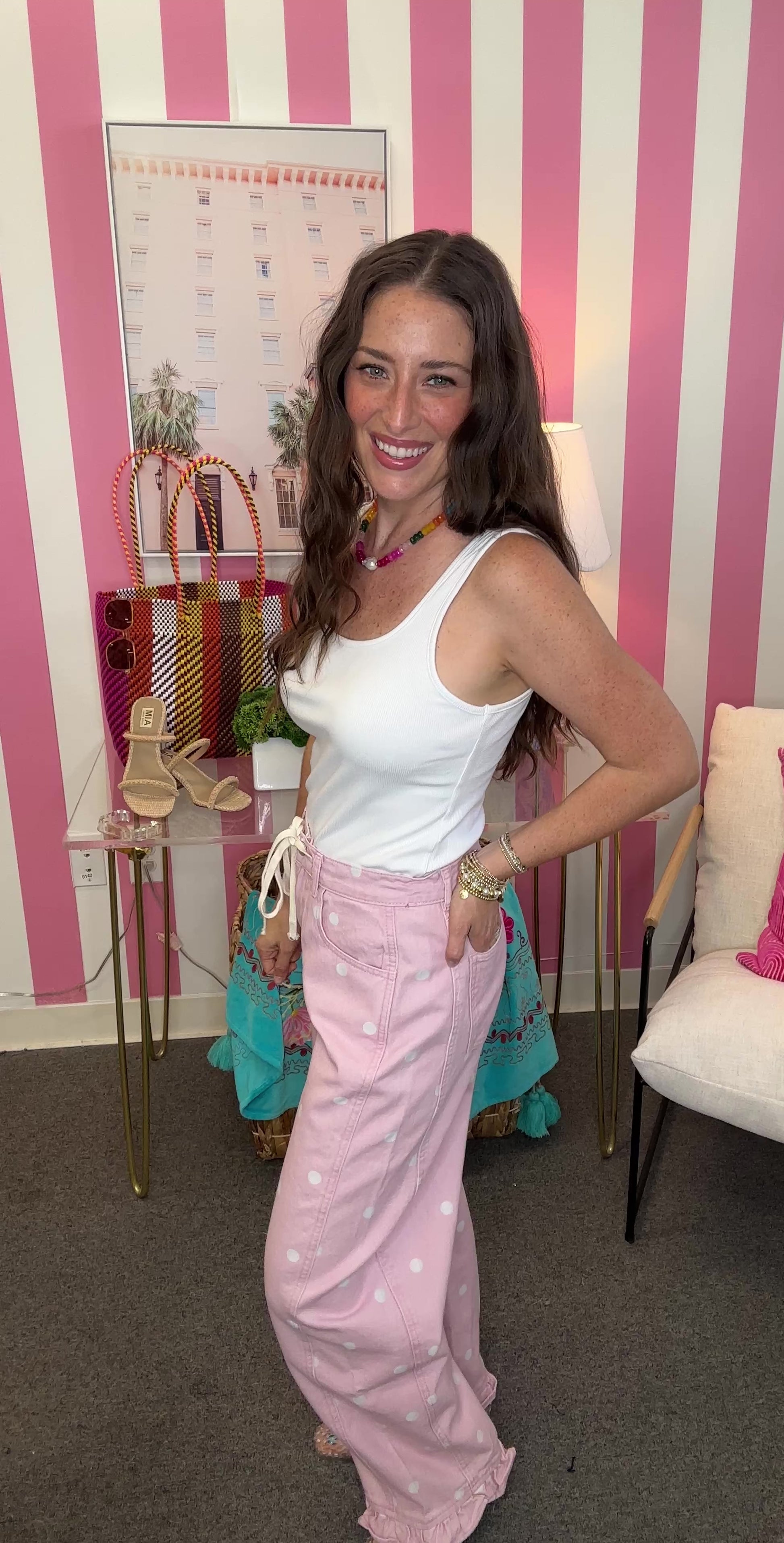 Woman in a white top and pink pants standing in a room with striped walls and furniture.