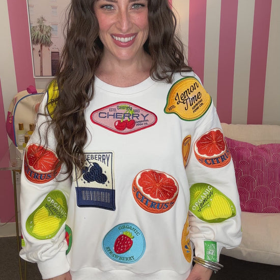 Woman wearing a white sweatshirt with colorful fruit patches in a room with pink and white striped wallpaper.