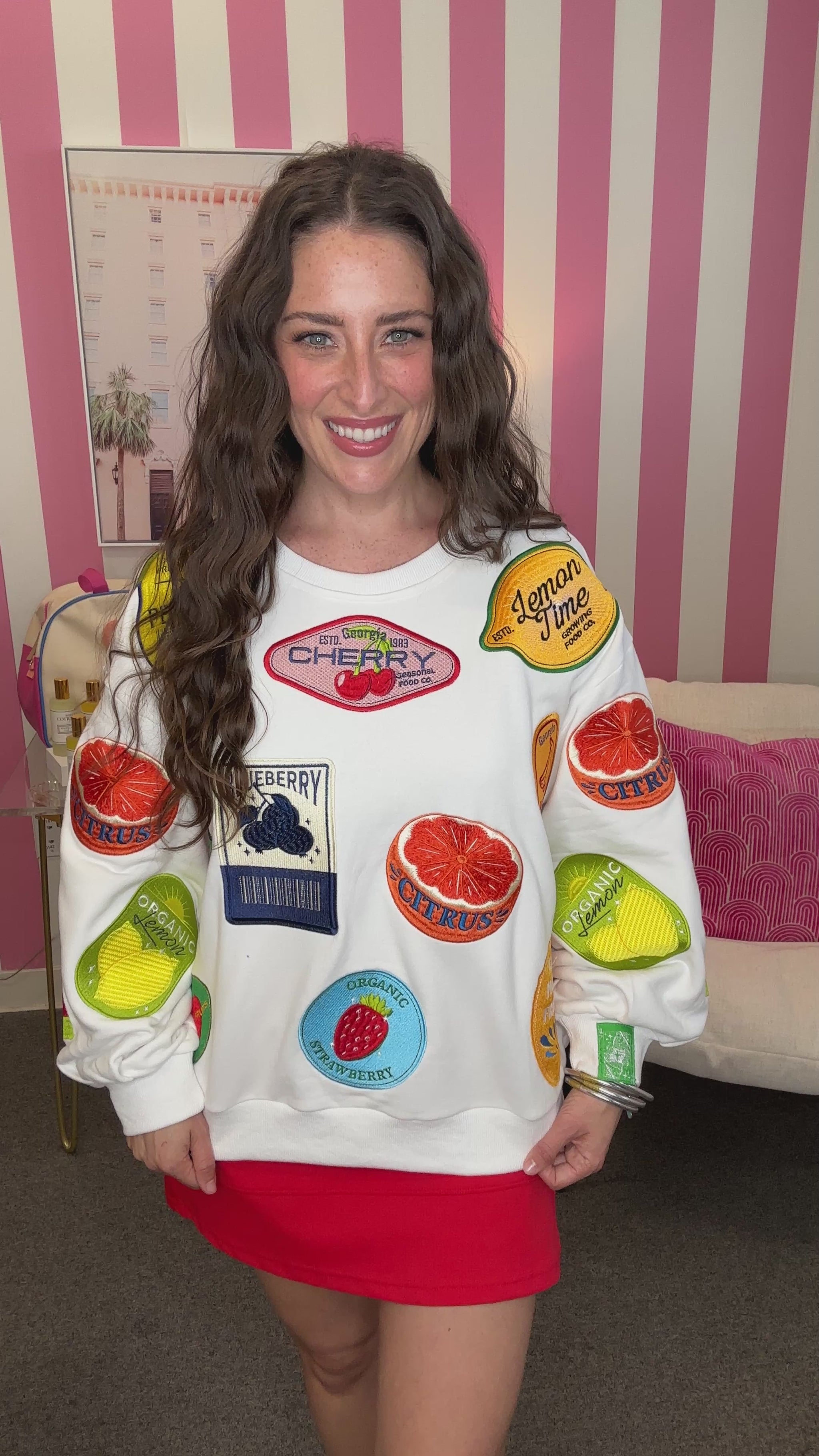 Woman wearing a white sweatshirt with colorful fruit patches in a room with pink and white striped wallpaper.