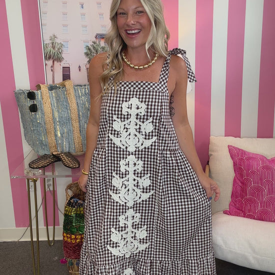 A woman modeling a black and white gingham maxi dress with white lace applique down the front, standing in front of a pink and white striped wall.
