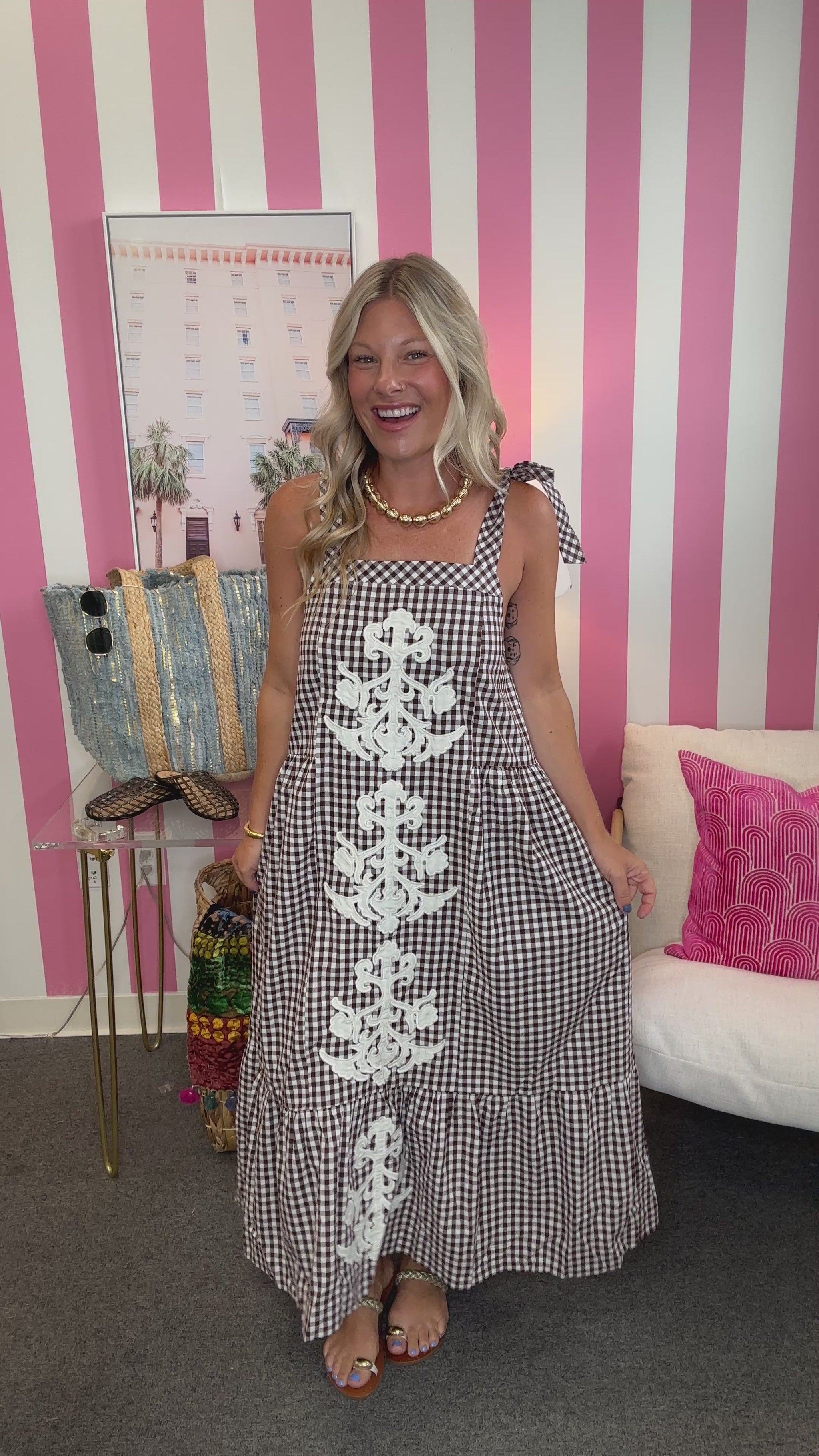 A woman modeling a black and white gingham maxi dress with white lace applique down the front, standing in front of a pink and white striped wall.